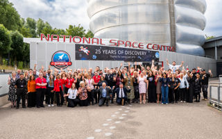 The employees of the National Space Centre gather in front of the building and cheer. Above them a sign reads '25 Years of Discovery'.