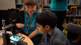 Child in IGNITE programme in a library with a Discovery team member looking at a meteorite under a microscope