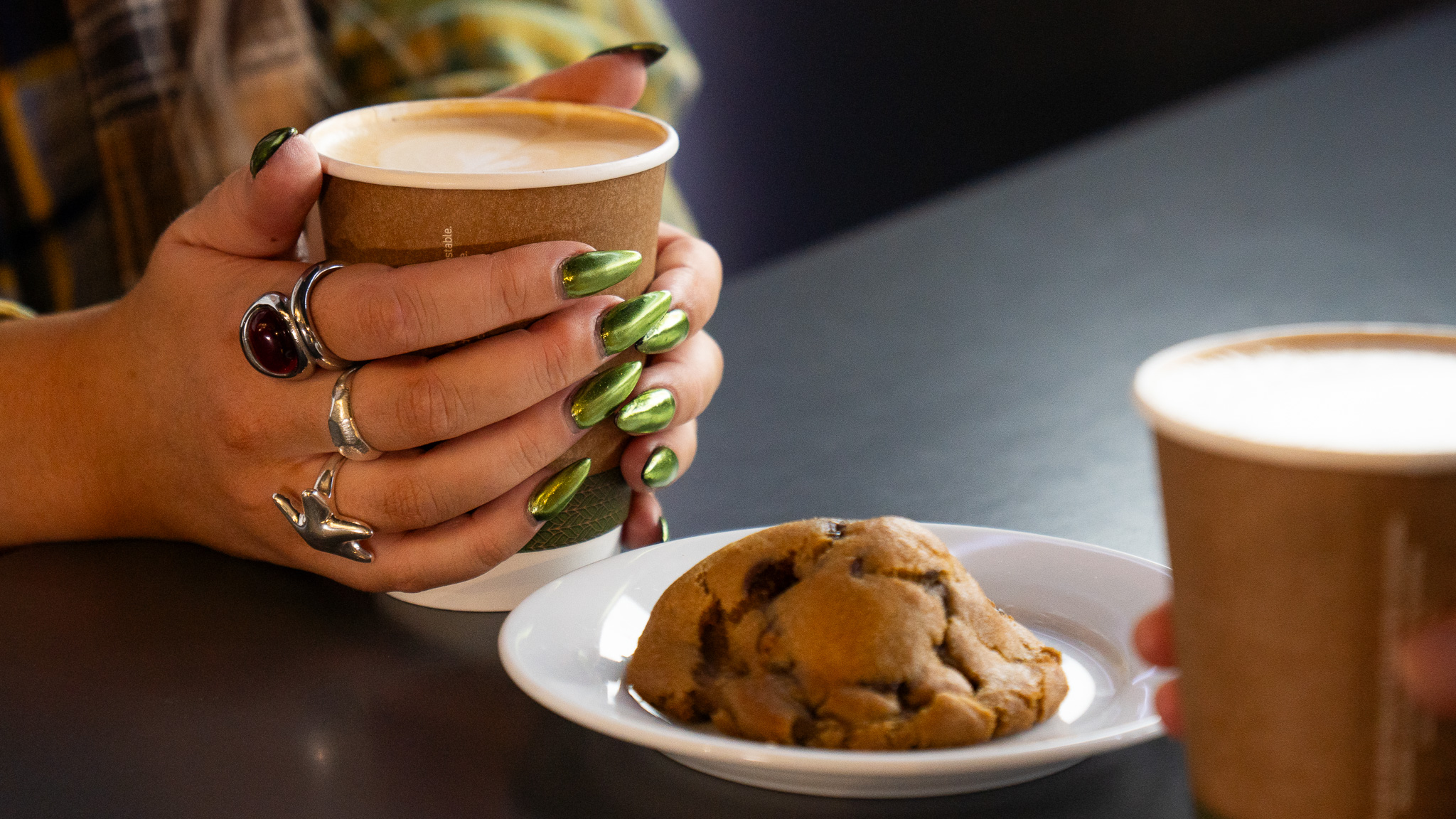 A woman sits holding a cup of coffee with a cookie in front of her. 